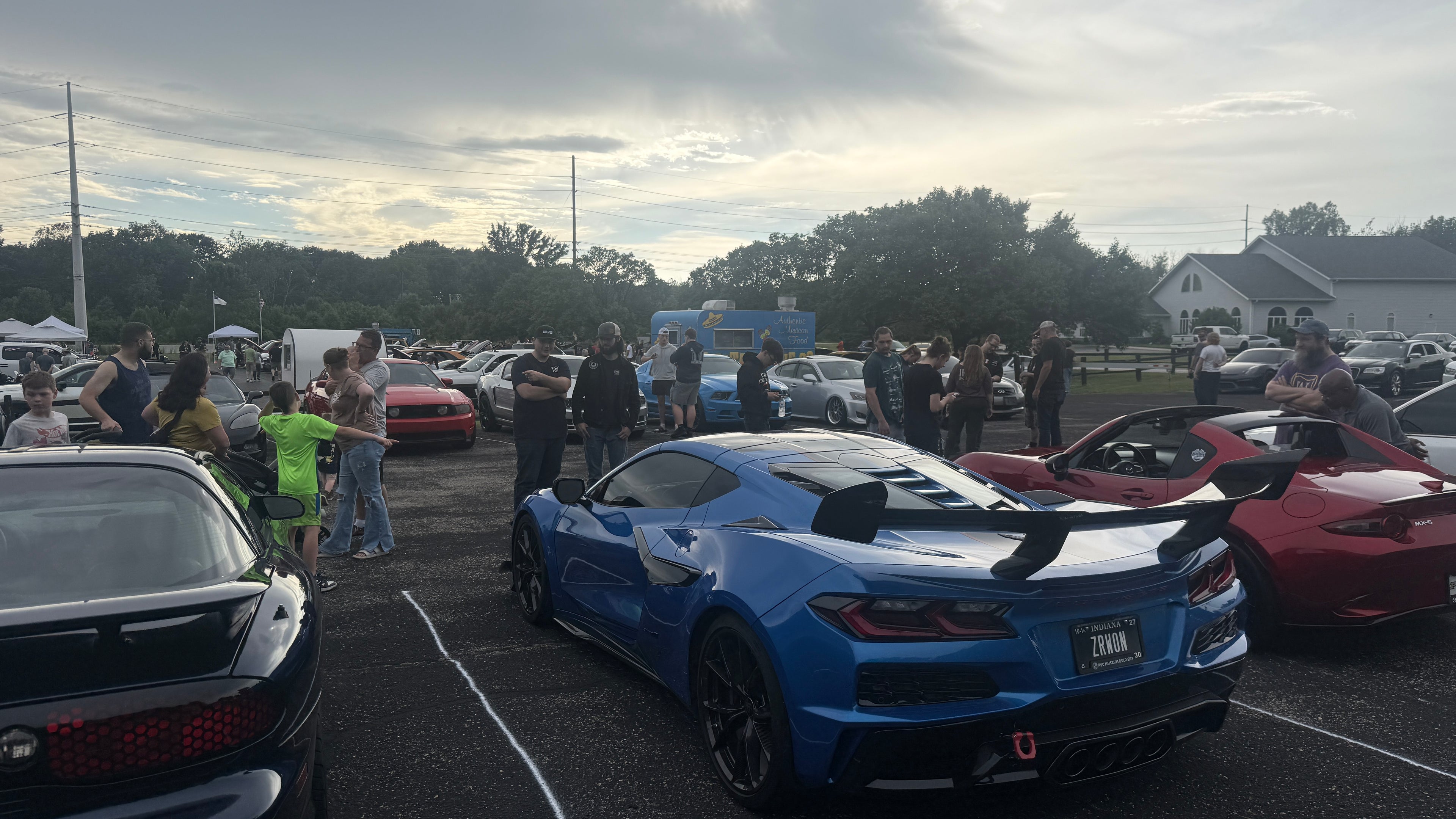 A blue sports car parked in a crowded outdoor car show or parking lot, with people and other cars visible around it.